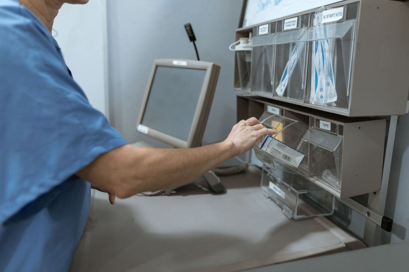 A nurse in blue scrubs takes supplies from a wall cabinet beside a computer screen.