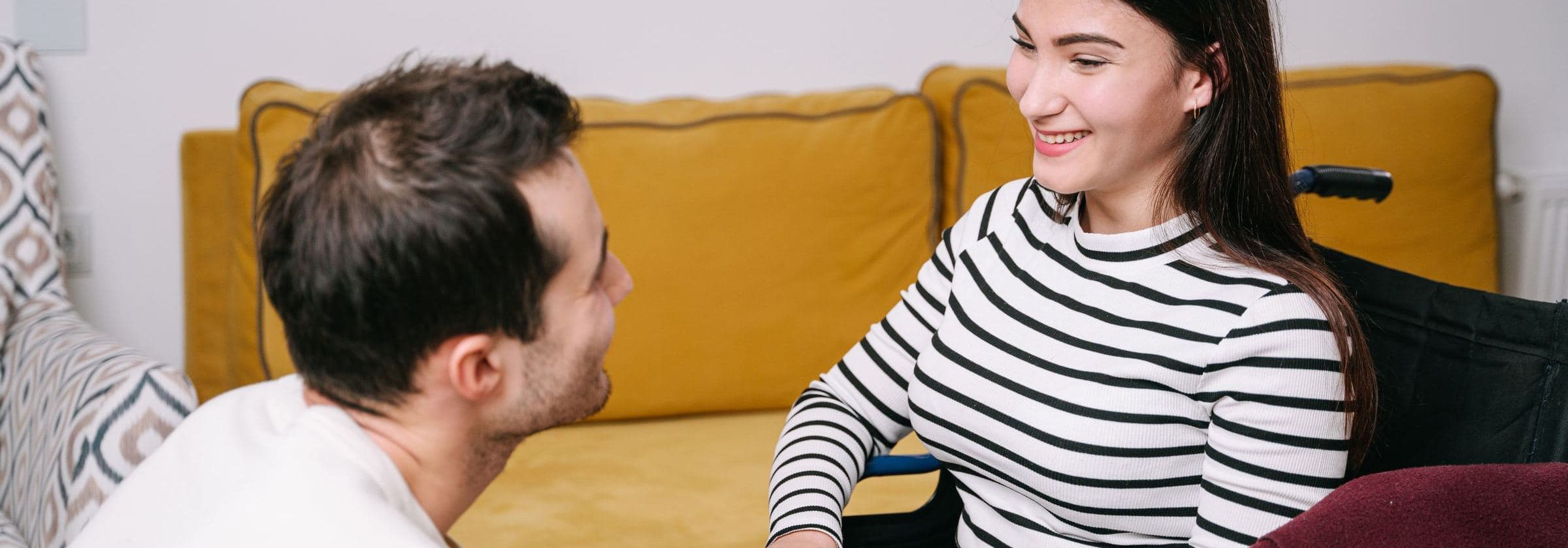 A care worker kneels to talk with a smiling young woman in a wheelchair beside a yellow sofa.