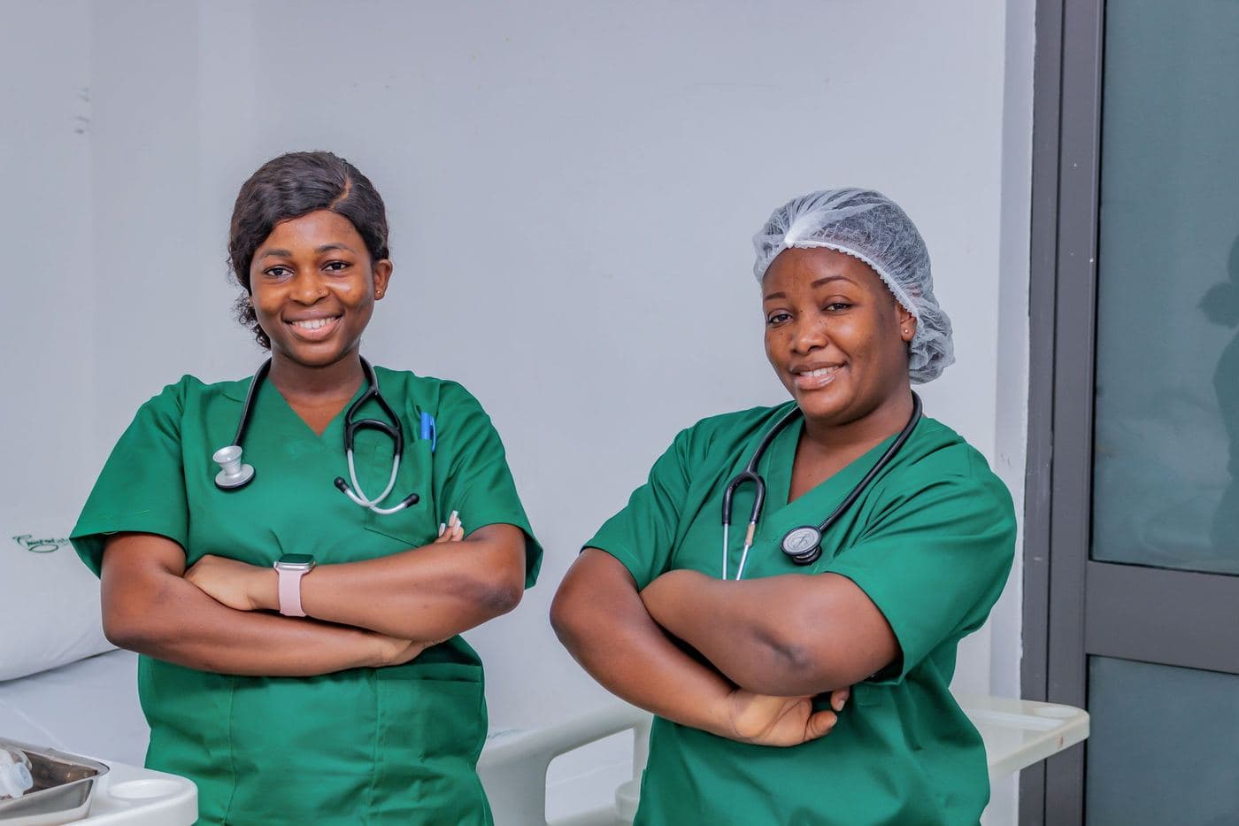 Two nurses in green scrubs stand side by side, arms folded, smiling.