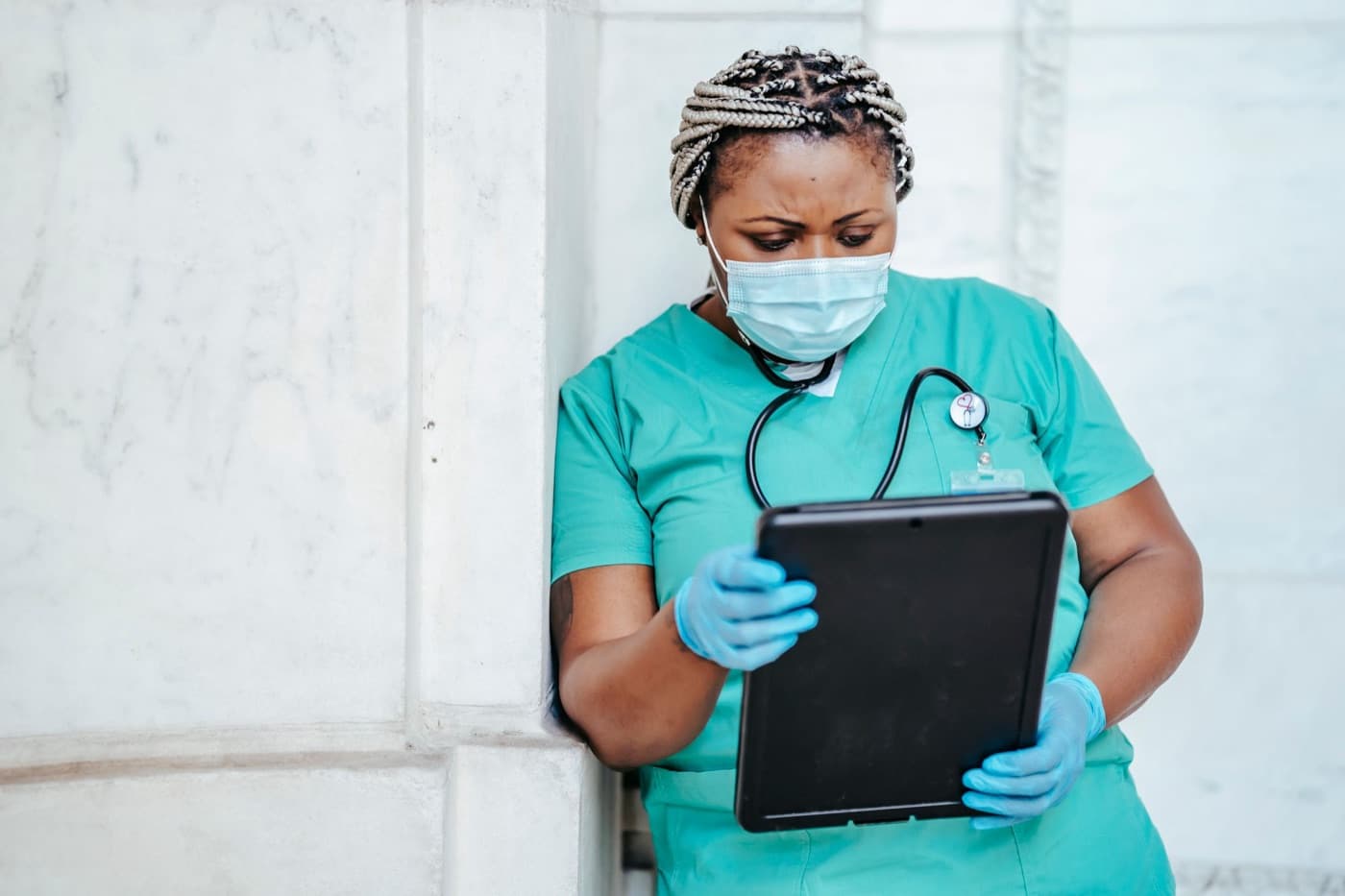 A nurse in scrubs holding a tablet and reading it, standing against a pale stone wall.
