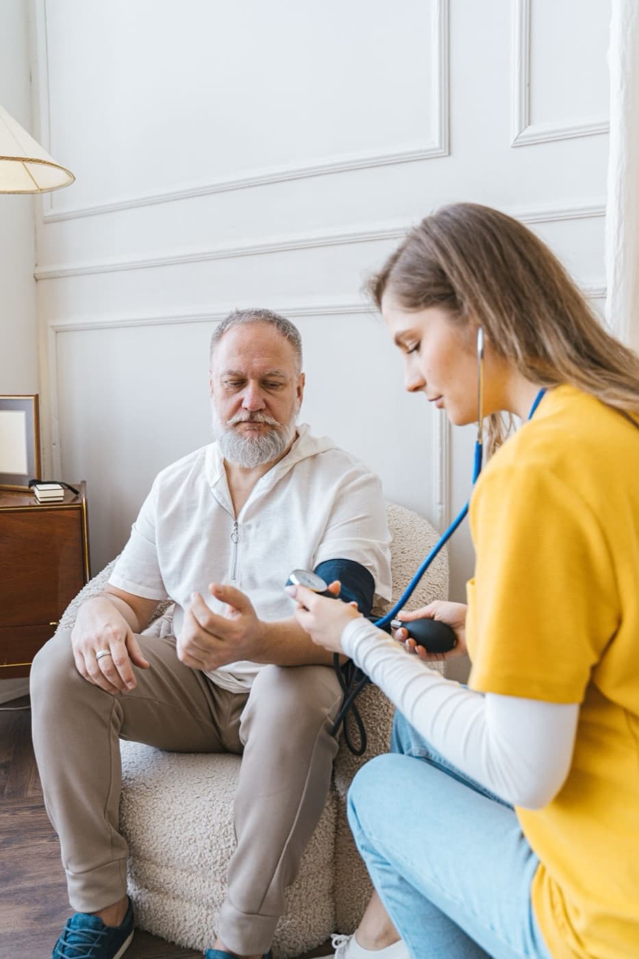 A home-care worker in a yellow top taking an older man's blood pressure in a sunlit sitting room.