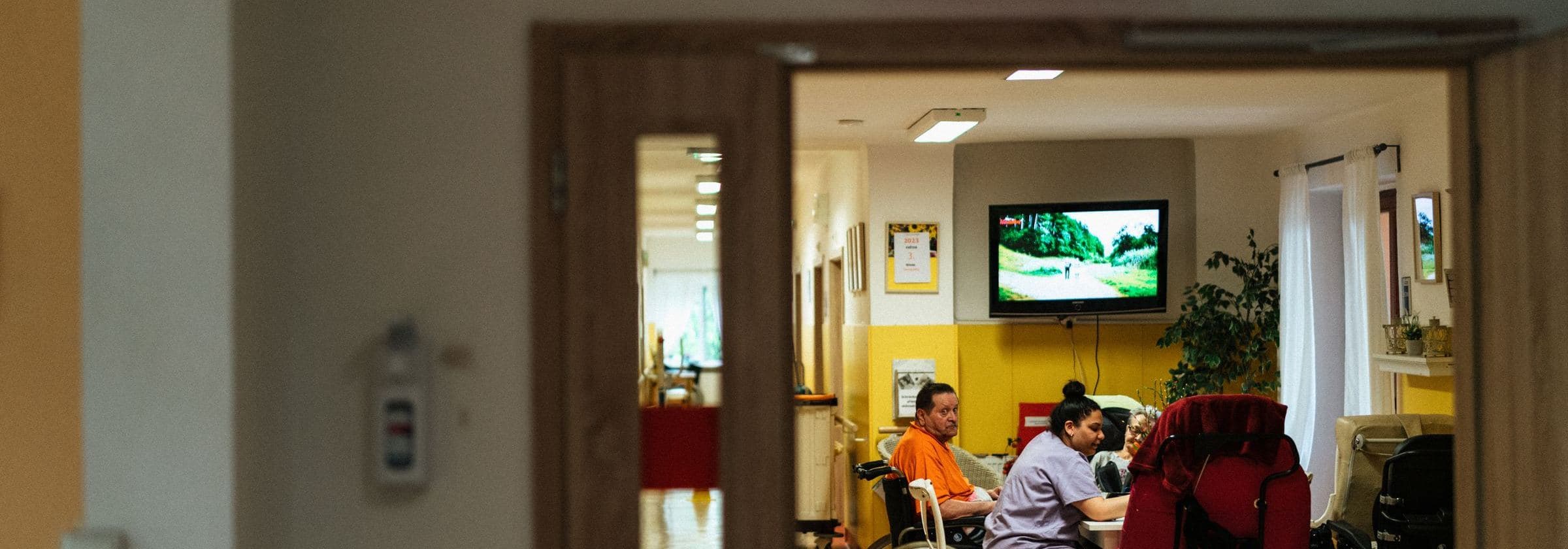 Through an open doorway, staff and residents in a residential care lounge with warm yellow walls.