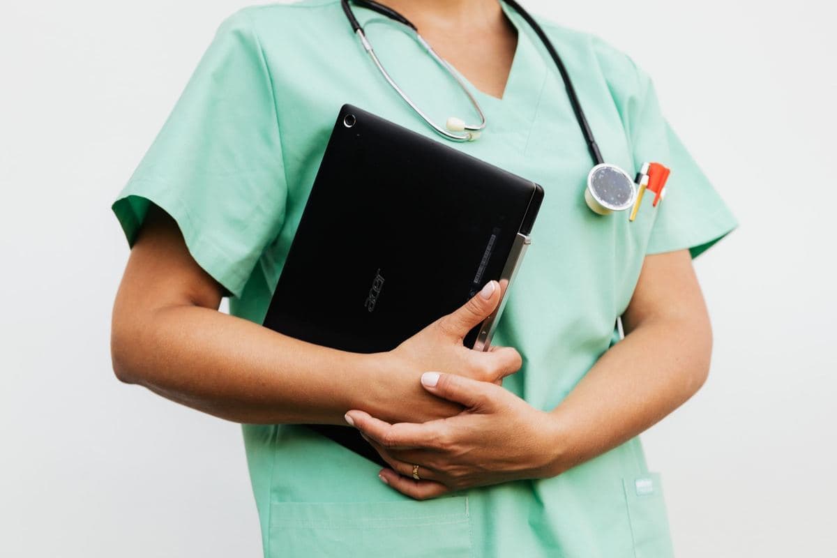 A nurse in green scrubs holds a tablet against her chest, a stethoscope around her neck.