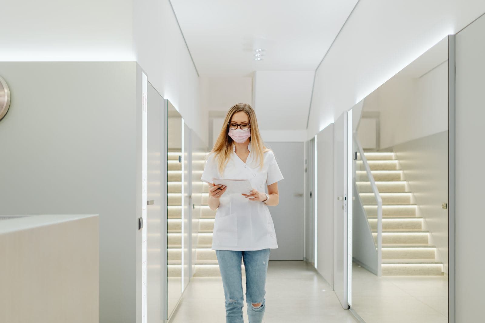 A nurse in a mask walks down a bright white hospital corridor carrying a tablet.