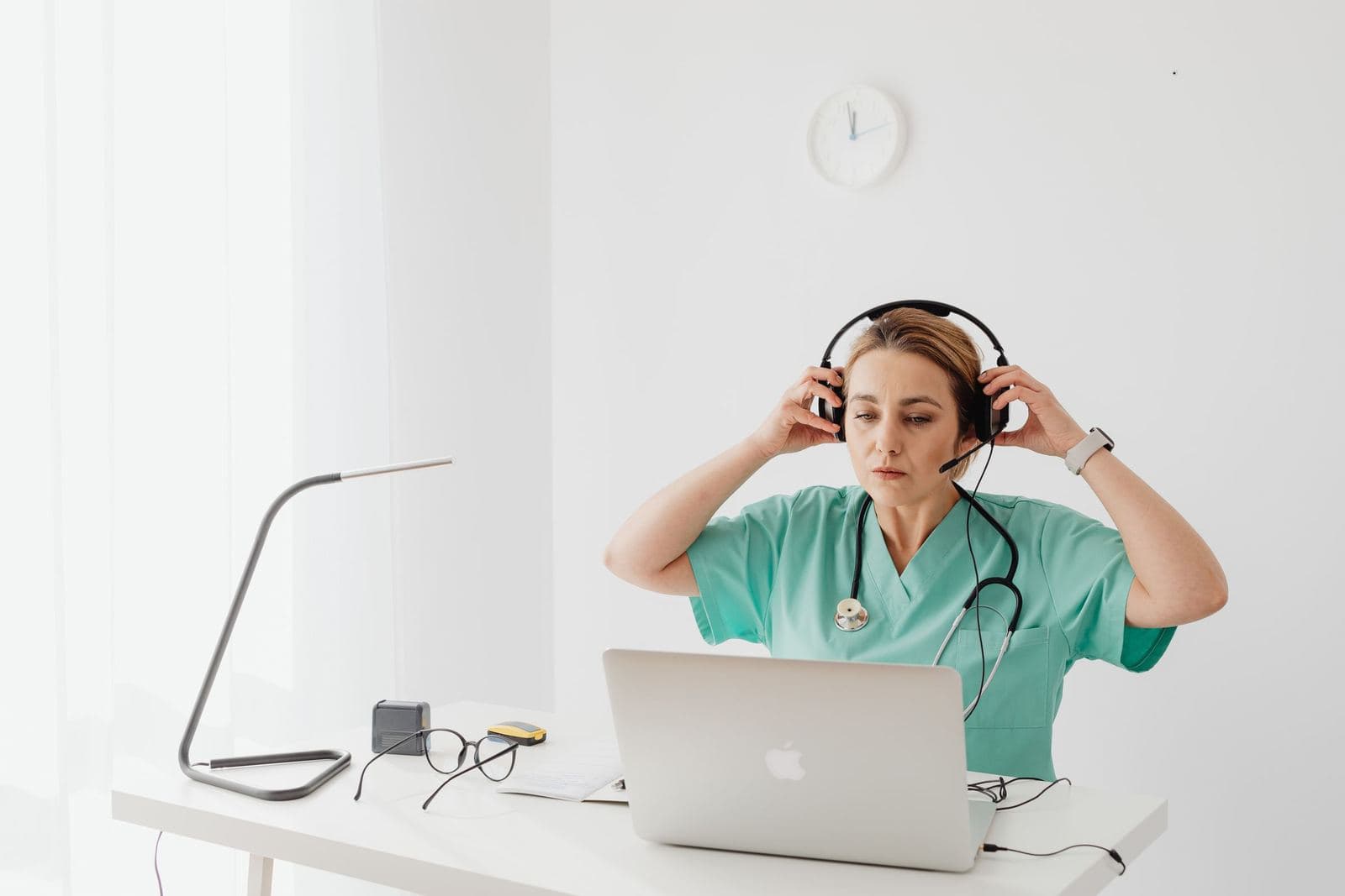 A nurse in green scrubs puts on a headset at a desk with a laptop.